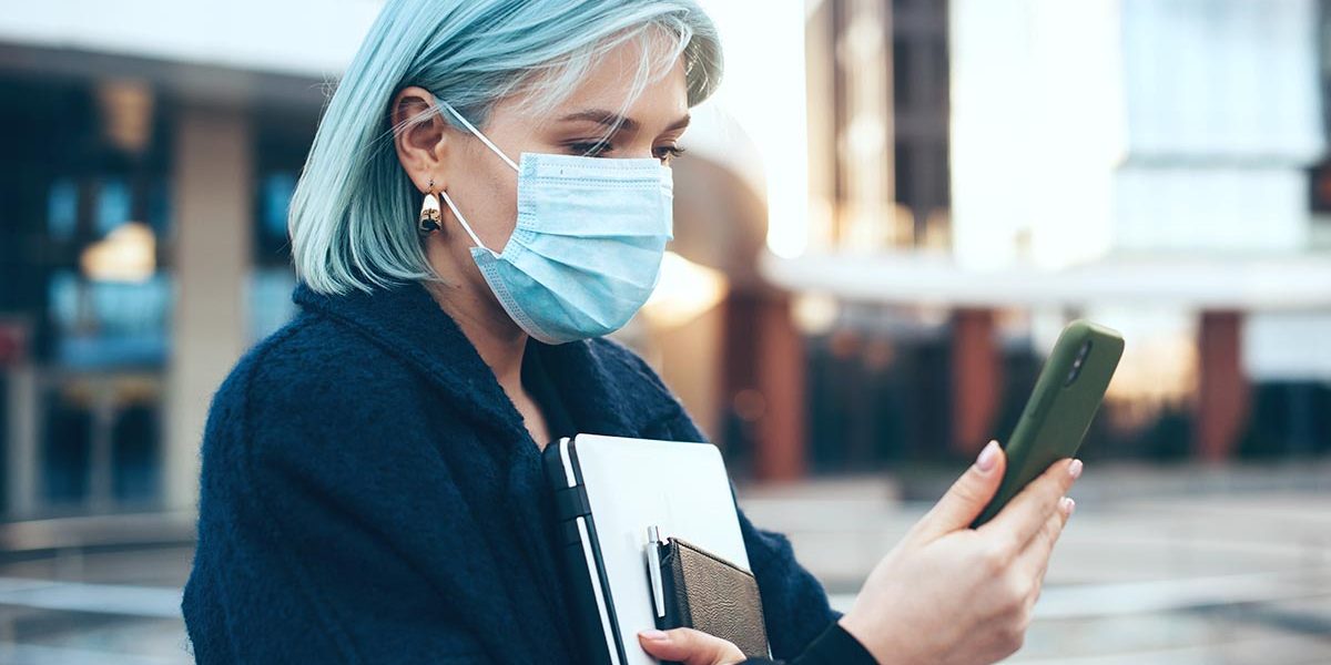Blue haired caucasian woman posing outside with a computer and phone while wearing a special anti virus mask